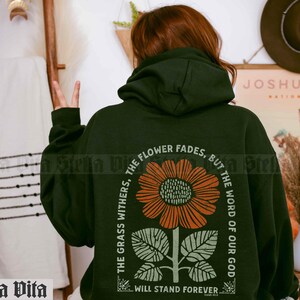 May include: A dark green hoodie with a floral graphic and the text "The grass withers, the flower fades, but the word of our God will stand forever."