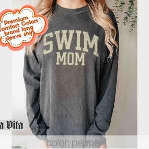 Puede incluir: Una camiseta negra de manga larga con el texto "SWIM MOM" en letras doradas. La camiseta est&aacute; hecha de una tela suave y c&oacute;moda.