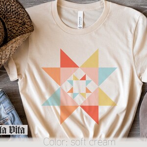 May include: A soft cream t-shirt with a colorful geometric star design. The star is made up of triangles in shades of pink, orange, yellow, and blue. The shirt is a simple, casual style with a crew neck and short sleeves. "Stella Vita" is written in black at the bottom left of the image. "Color: soft cream" is written in black at the bottom right of the image.
