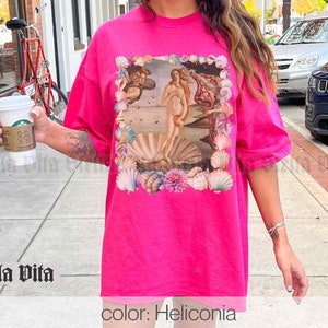 Puede incluir: Una camiseta rosa de gran tama&ntilde;o con un estampado gr&aacute;fico de la pintura El nacimiento de Venus de Sandro Botticelli. La impresi&oacute;n est&aacute; rodeada por un borde de conchas marinas. El texto "color: Heliconia" est&aacute; impreso en la parte inferior de la imagen.