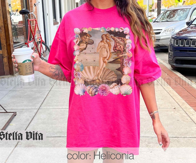 Puede incluir: Una camiseta rosa de gran tama&ntilde;o con un estampado gr&aacute;fico de la pintura El nacimiento de Venus de Sandro Botticelli. La impresi&oacute;n est&aacute; rodeada por un borde de conchas marinas. El texto "color: Heliconia" est&aacute; impreso en la parte inferior de la imagen.
