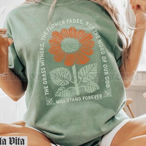 May include: A green t-shirt with a floral design and the text "The flower fades, but the word of our God, the grass withers, will stand forever".