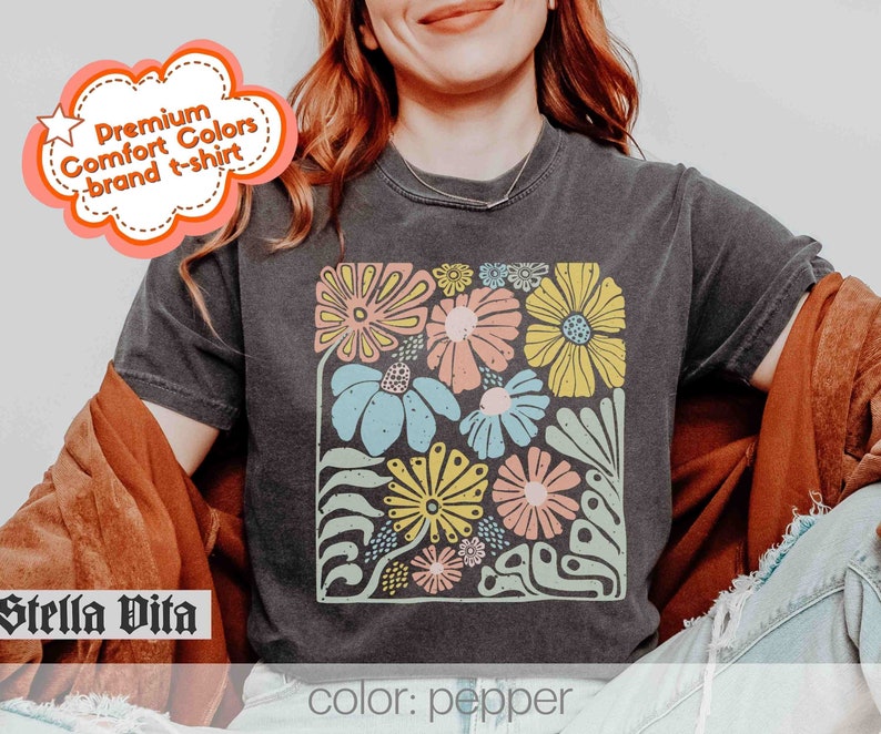 Peut inclure: Un t-shirt gris fonc&eacute; avec un imprim&eacute; floral color&eacute;. L'imprim&eacute; pr&eacute;sente une vari&eacute;t&eacute; de fleurs dans des tons de rose, jaune, bleu et vert. Le texte "Stella Vita" est visible dans le coin inf&eacute;rieur gauche de l'image. Le texte "color: pepper" est visible dans le coin inf&eacute;rieur droit de l'image.