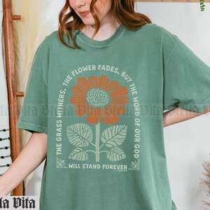 May include: Green t-shirt with a floral design and the text "The grass withers, the flower fades, but the word of our God will stand forever".
