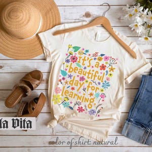 May include: A white t-shirt with a floral design and the text "It's a beautiful day for learning." The shirt is on a wooden surface with a straw hat, sandals, and blue jeans.