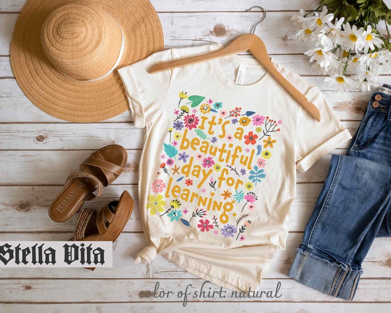 May include: A white t-shirt with a floral design and the text "It's a beautiful day for learning." The shirt is on a wooden surface with a straw hat, sandals, and blue jeans.
