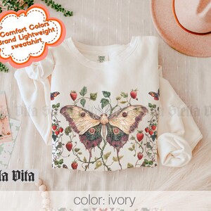 May include: A white sweatshirt with a butterfly and floral print. The text "Comfort Colors Brand Lightweight Sweatshirt" is on a pink cloud shape. The color of the sweatshirt is ivory.