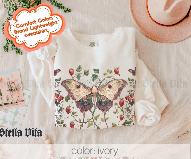 May include: A white sweatshirt with a butterfly and floral print. The text "Comfort Colors Brand Lightweight Sweatshirt" is on a pink cloud shape. The color of the sweatshirt is ivory.