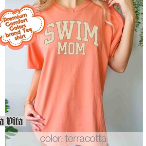 Puede incluir: Camiseta de color terracota con el texto "Swim Mom" en color crema.