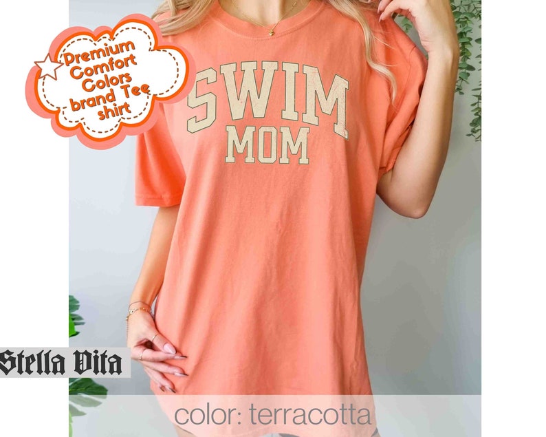 Puede incluir: Camiseta de color terracota con el texto "Swim Mom" en color crema.