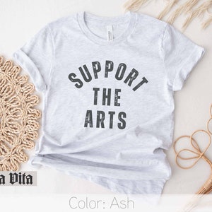 May include: A light gray t-shirt with the text "SUPPORT THE ARTS" printed in black. The text is in a distressed font style.