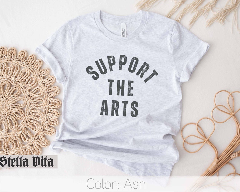 May include: A light gray t-shirt with the text "SUPPORT THE ARTS" printed in black. The text is in a distressed font style.