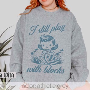 K&ouml;nnte beinhalten: Ein graues Sweatshirt mit dem Text "I still play with blocks" und einer Cartoon-Illustration eines M&auml;dchens, das mit Bl&ouml;cken spielt.