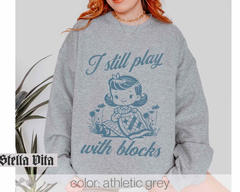 K&ouml;nnte beinhalten: Ein graues Sweatshirt mit dem Text "I still play with blocks" und einer Cartoon-Illustration eines M&auml;dchens, das mit Bl&ouml;cken spielt.
