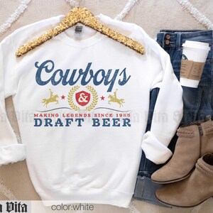 May include: White sweatshirt with the word "Cowboys" in blue script, and "DRAFT BEER" in blue block letters. The design includes a gold emblem with a rider on a horse and the text "MAKING LEGENDS SINCE 1985". The sweatshirt is displayed with jeans and boots.
