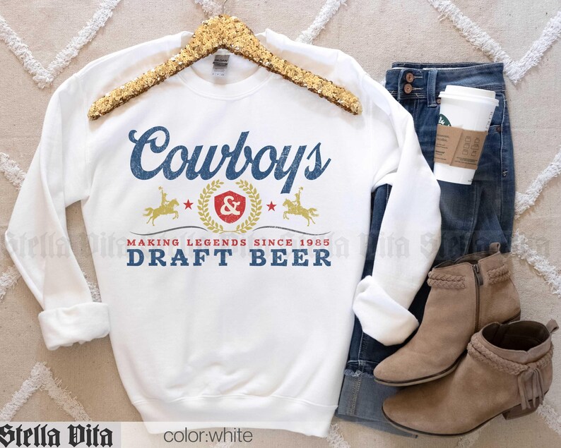 May include: White sweatshirt with the word "Cowboys" in blue script, and "DRAFT BEER" in blue block letters. The design includes a gold emblem with a rider on a horse and the text "MAKING LEGENDS SINCE 1985". The sweatshirt is displayed with jeans and boots.