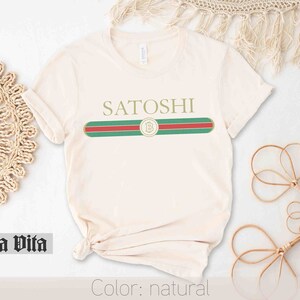 May include: A natural colored t-shirt with a green, red, and white stripe across the chest. The stripe has a gold circle with a "B" inside. The text "SATOSHI" is above the stripe.