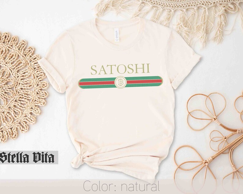 May include: A natural colored t-shirt with a green, red, and white stripe across the chest. The stripe has a gold circle with a "B" inside. The text "SATOSHI" is above the stripe.