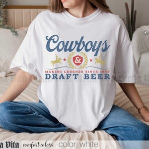 May include: White t-shirt with a blue and gold graphic that reads "Cowboys" above a crest and the words "Draft Beer". The shirt also has the text "Making Legends Since 1985". The shirt is white.