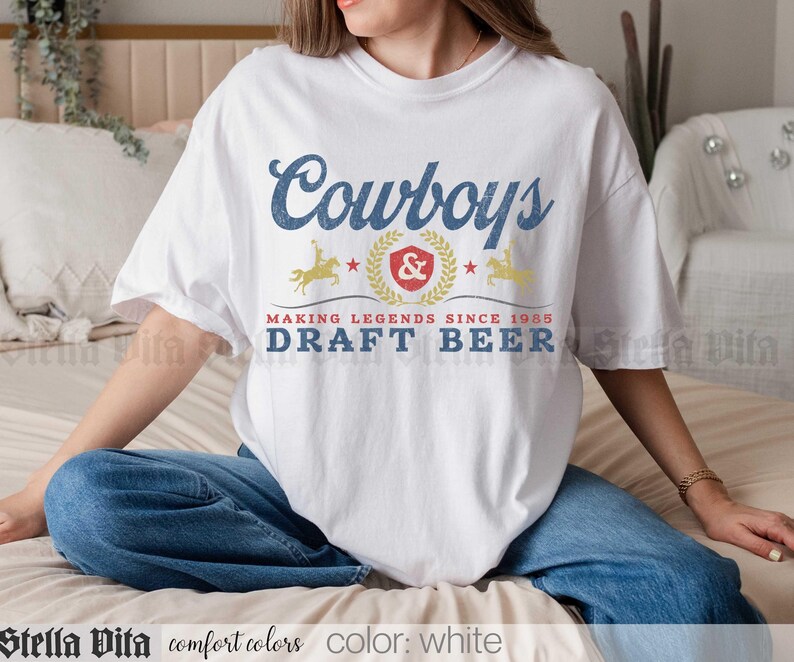 May include: White t-shirt with a blue and gold graphic that reads "Cowboys" above a crest and the words "Draft Beer". The shirt also has the text "Making Legends Since 1985". The shirt is white.