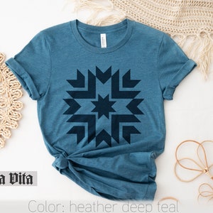 May include: A teal blue t-shirt with a dark blue geometric starburst design. The text "Stella Vita" is printed on the bottom left corner of the image. The text "Color: heather deep teal" is printed on the bottom right corner of the image.