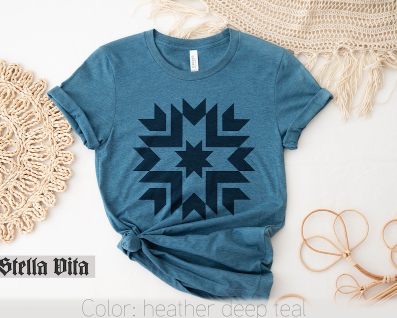 May include: A teal blue t-shirt with a dark blue geometric starburst design. The text "Stella Vita" is printed on the bottom left corner of the image. The text "Color: heather deep teal" is printed on the bottom right corner of the image.