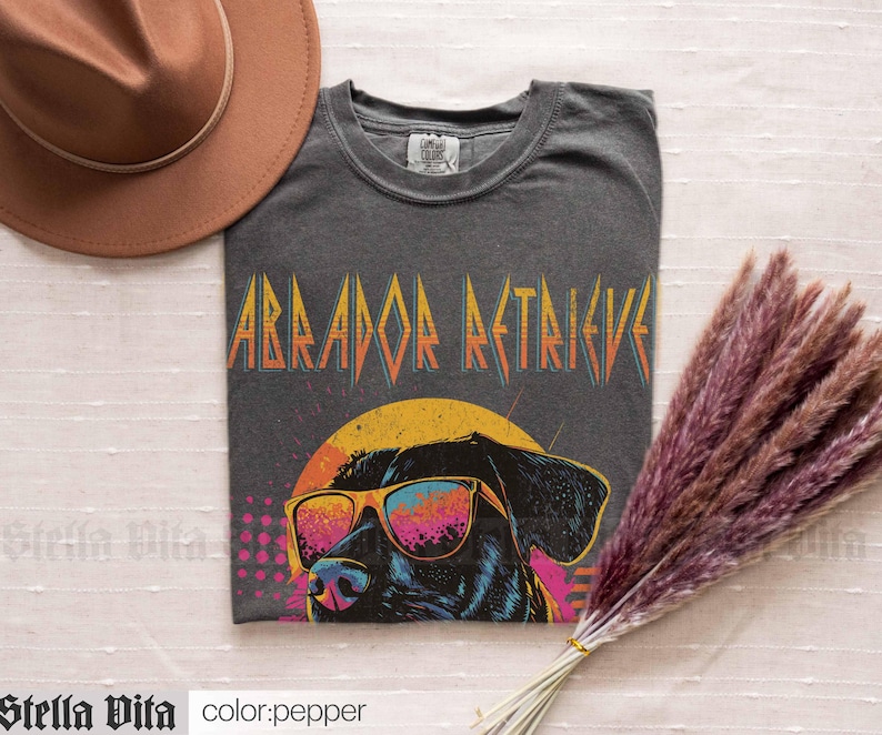 Puede incluir: Una camiseta gris oscuro con un gr&aacute;fico retro de un Labrador Retriever con gafas de sol. El texto "Labrador Retriever" est&aacute; impreso en una fuente audaz y colorida.