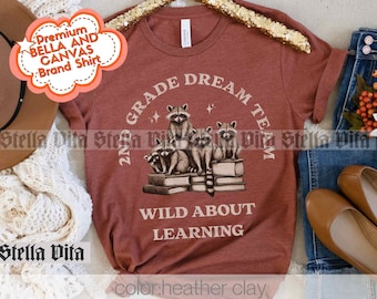 Puede incluir: Una camiseta de color óxido con un gráfico de mapaches leyendo libros. El texto de la camiseta dice "2nd Grade Dream Team Wild About Learning".