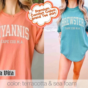 May include: Two t-shirts with a vintage college style design. The first t-shirt is terracotta with the text "HYANNIS CAPE COD M.A." The second t-shirt is seafoam green with the text "BREWSTER CAPE COD M.A."