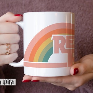 May include: White ceramic mug with a rainbow design and the word "RELAX" in pink letters. The mug is being held by a hand.
