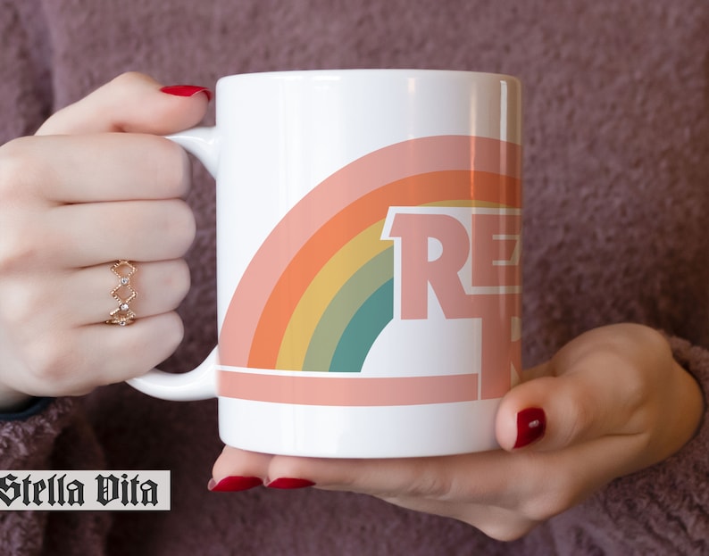 May include: White ceramic mug with a rainbow design and the word "RELAX" in pink letters. The mug is being held by a hand.