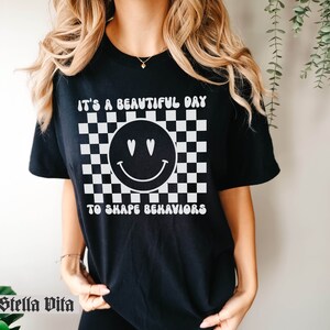 May include: Black t-shirt with a white checkered background, a smiley face with hearts for eyes, and the text "It's a beautiful day to shape behaviors".