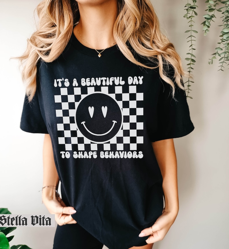 May include: Black t-shirt with a white checkered background, a smiley face with hearts for eyes, and the text "It's a beautiful day to shape behaviors".