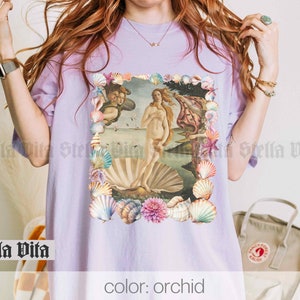 Puede incluir: Una camiseta de color lavanda con un estampado gr&aacute;fico de la pintura El nacimiento de Venus rodeada de conchas. El texto "color: orqu&iacute;dea" est&aacute; impreso en la parte inferior de la imagen.