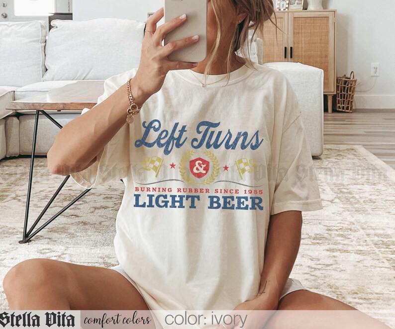 May include: Ivory-colored t-shirt with the text "Left Turns" in blue and yellow. The shirt also features the text "BURNING RUBBER SINCE 1985 LIGHT BEER". The shirt is part of the "comfort colors" collection.