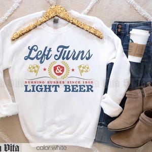 May include: White sweatshirt with the text "Left Turns Light Beer" in blue and gold lettering. The design includes racing flags and a laurel wreath. The sweatshirt is displayed with blue jeans, tan boots, and a coffee cup.