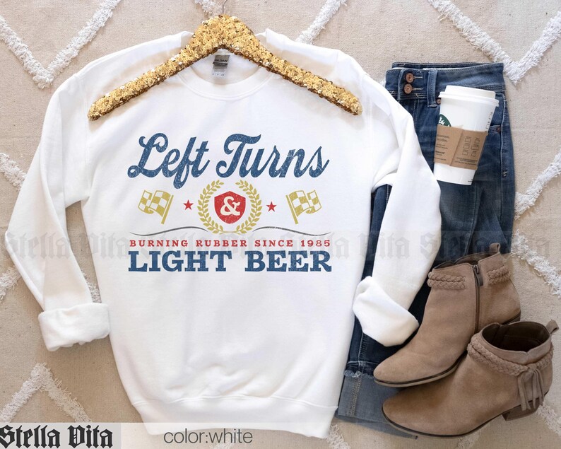 May include: White sweatshirt with the text "Left Turns Light Beer" in blue and gold lettering. The design includes racing flags and a laurel wreath. The sweatshirt is displayed with blue jeans, tan boots, and a coffee cup.
