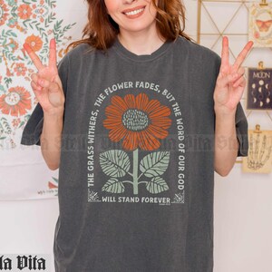 May include: A dark gray t-shirt with a graphic of a sunflower and the text "The grass withers, the flower fades, but the word of our God will stand forever."
