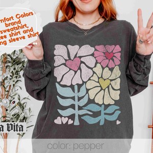May include: A black long-sleeved sweatshirt featuring a colourful floral design and the text "Comfort Colors brand sweatshirt, tee shirt and long sleeve shirt" and "Stella Vita" at the bottom. The text "color: pepper" is also visible.