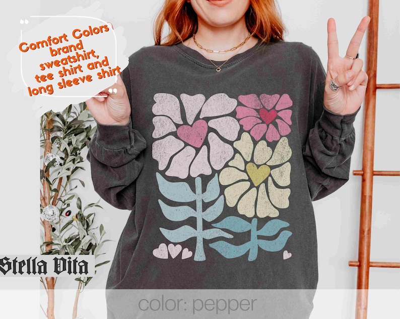 May include: A black long-sleeved sweatshirt featuring a colourful floral design and the text "Comfort Colors brand sweatshirt, tee shirt and long sleeve shirt" and "Stella Vita" at the bottom. The text "color: pepper" is also visible.