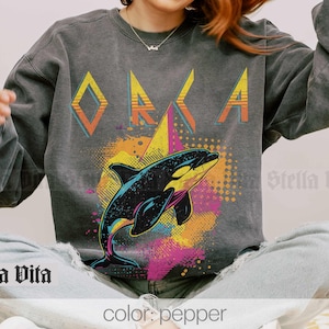 May include: A grey sweatshirt with a retro graphic design featuring an orca whale and the word "ORCA" in a bold, geometric font. The design is set against a colourful background with geometric shapes and a dotted pattern.