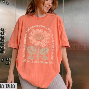 May include: A coral orange t-shirt with a floral graphic and the text "The grass withers, the flower fades, but the word of our God will stand forever".