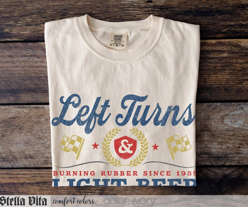 May include: Ivory-colored t-shirt with the text "Left Turns" in blue script. Below the text is a graphic with a shield, laurel wreath, and checkered flags. The shirt also has the text "BURNING RUBBER SINCE 1985 LIGHT BEER".