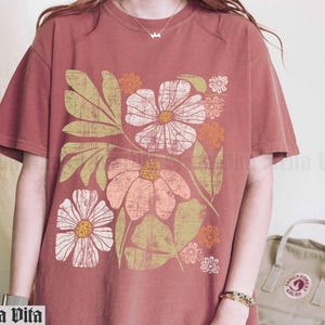 May include: A rust-colored t-shirt with a floral print featuring pink, white, and orange flowers. The flowers are stylized and have a retro feel.