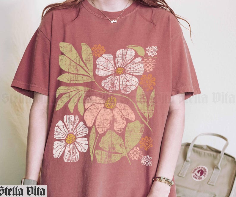 May include: A rust-colored t-shirt with a floral print featuring pink, white, and orange flowers. The flowers are stylized and have a retro feel.
