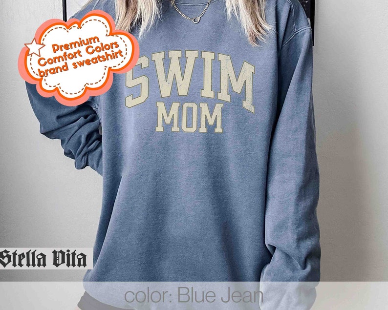 Puede incluir: Sudadera azul vaquero con el texto "SWIM MOM" en una fuente de estilo universitario vintage.