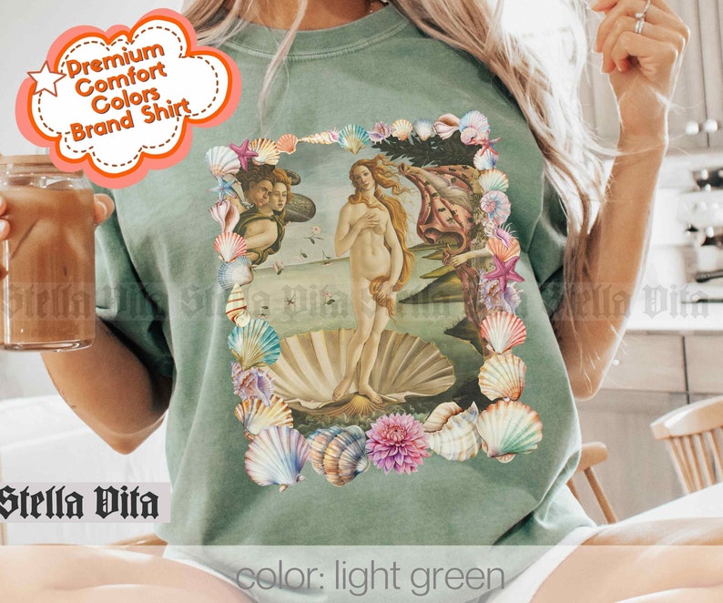 Puede incluir: Una camiseta verde claro con un estampado gr&aacute;fico de un borde de conchas que rodea una pintura de una mujer de pie sobre una concha. El texto "color: light green" est&aacute; impreso en la parte inferior de la imagen.