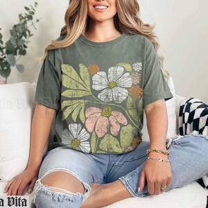 May include: A green t-shirt with a floral print featuring pink, white, and yellow flowers. The shirt has a relaxed fit and a crew neck.