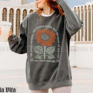 May include: A dark gray sweatshirt with a graphic of a sunflower and the text "The grass withers, the flower fades, but the word of our God will stand forever".