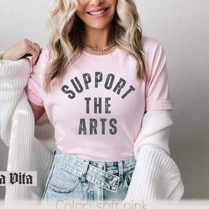 May include: A pink t-shirt with the text "Support the Arts" in gray lettering. The shirt is being worn by a person with long blonde hair.